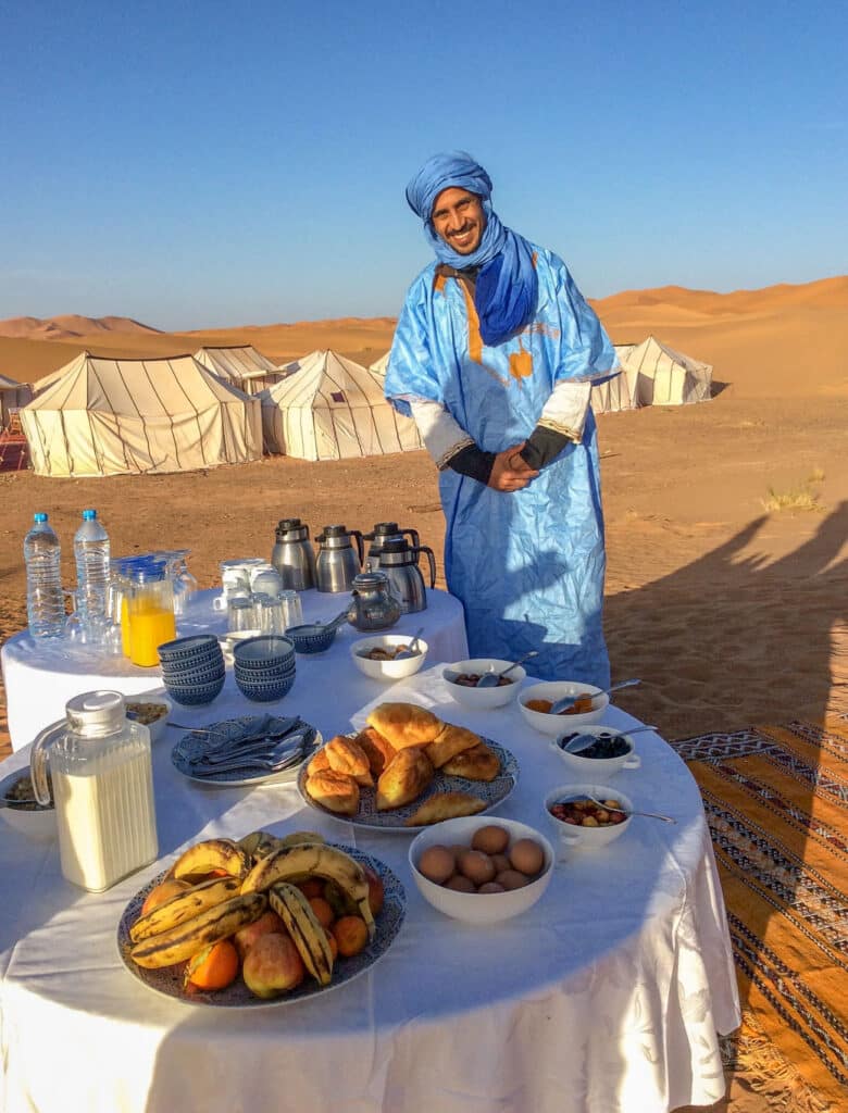 Smiling host in blue robe and turban stands in front of a table laden with fruits and pasteries in the Sahara Desert.