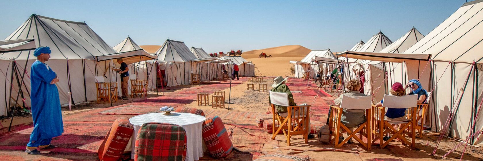 Travelers lounge among a dozen white canvas tents set up in Morocco’s Sahara Desert with colorful carpets and futons.