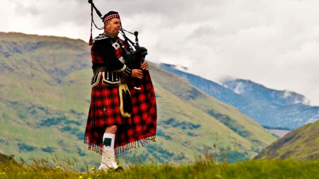 A piper plays the bagpipes on a hillside in traditional Scottish dress including kilt.