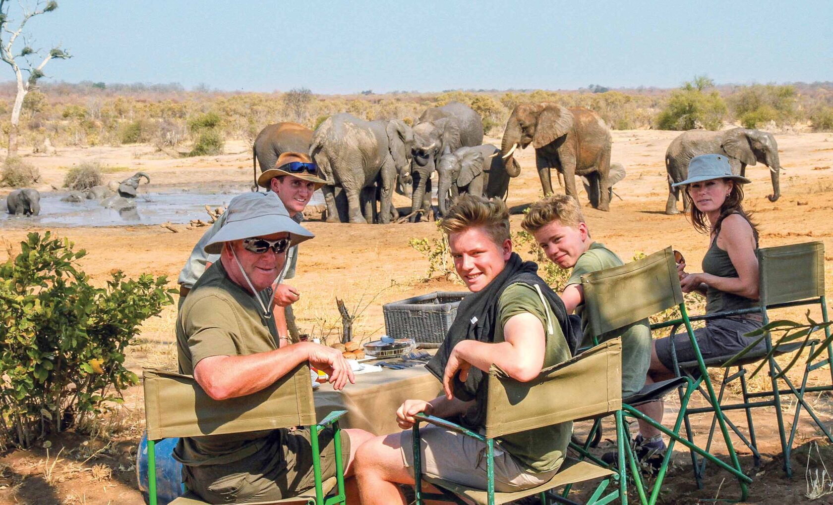 Group of travelers on safari in Zimbabwe having snacks near herd of elephants.